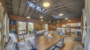 A spacious living area with a wooden dining table, comfortable seating, a kitchen area, and large windows providing natural light. - Bonners Ferry Cabin w/ Wraparound Deck & Views! (Bonners Ferry)