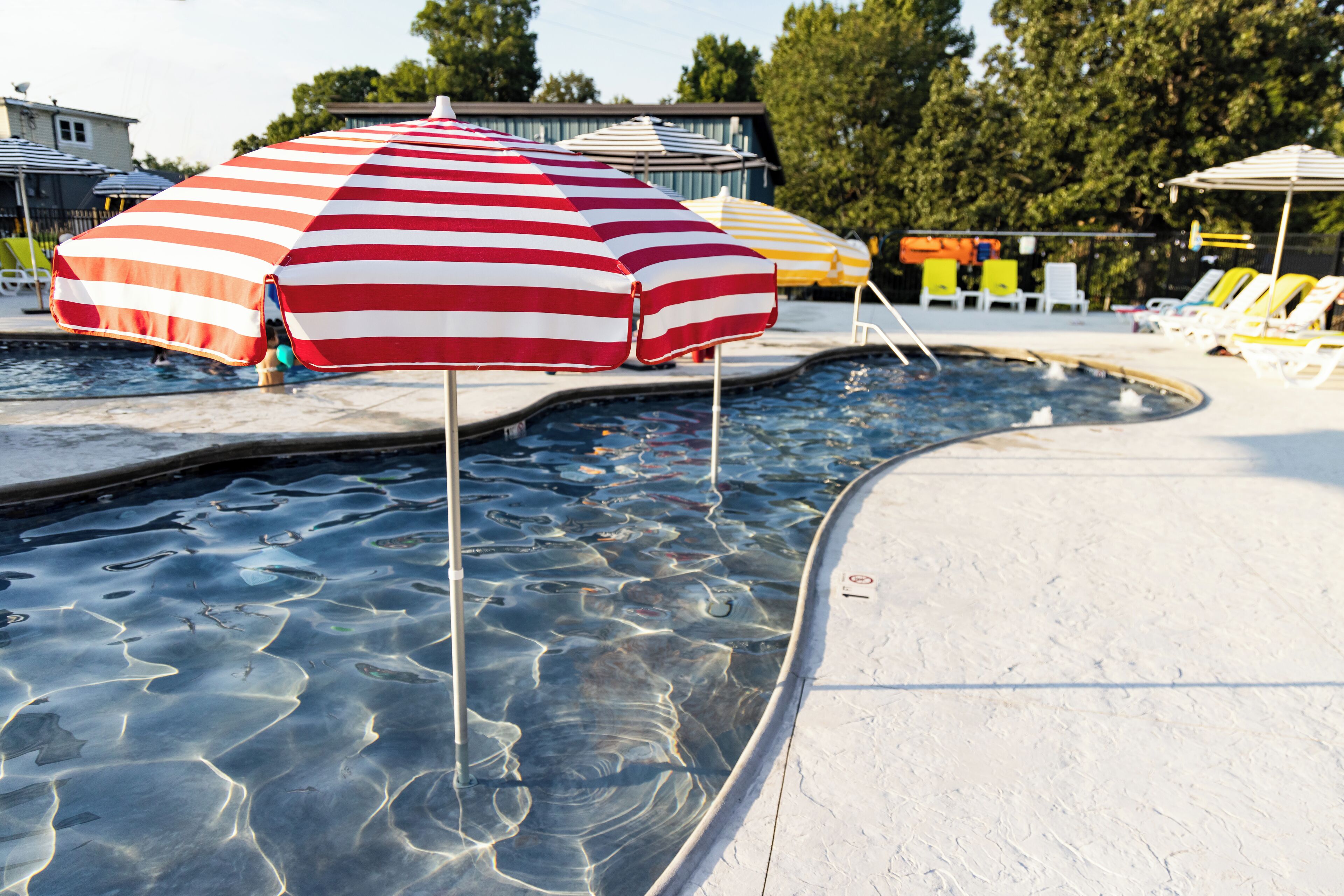 A swimming pool with a red and white striped umbrella, lounge chairs, and a clear water surface.