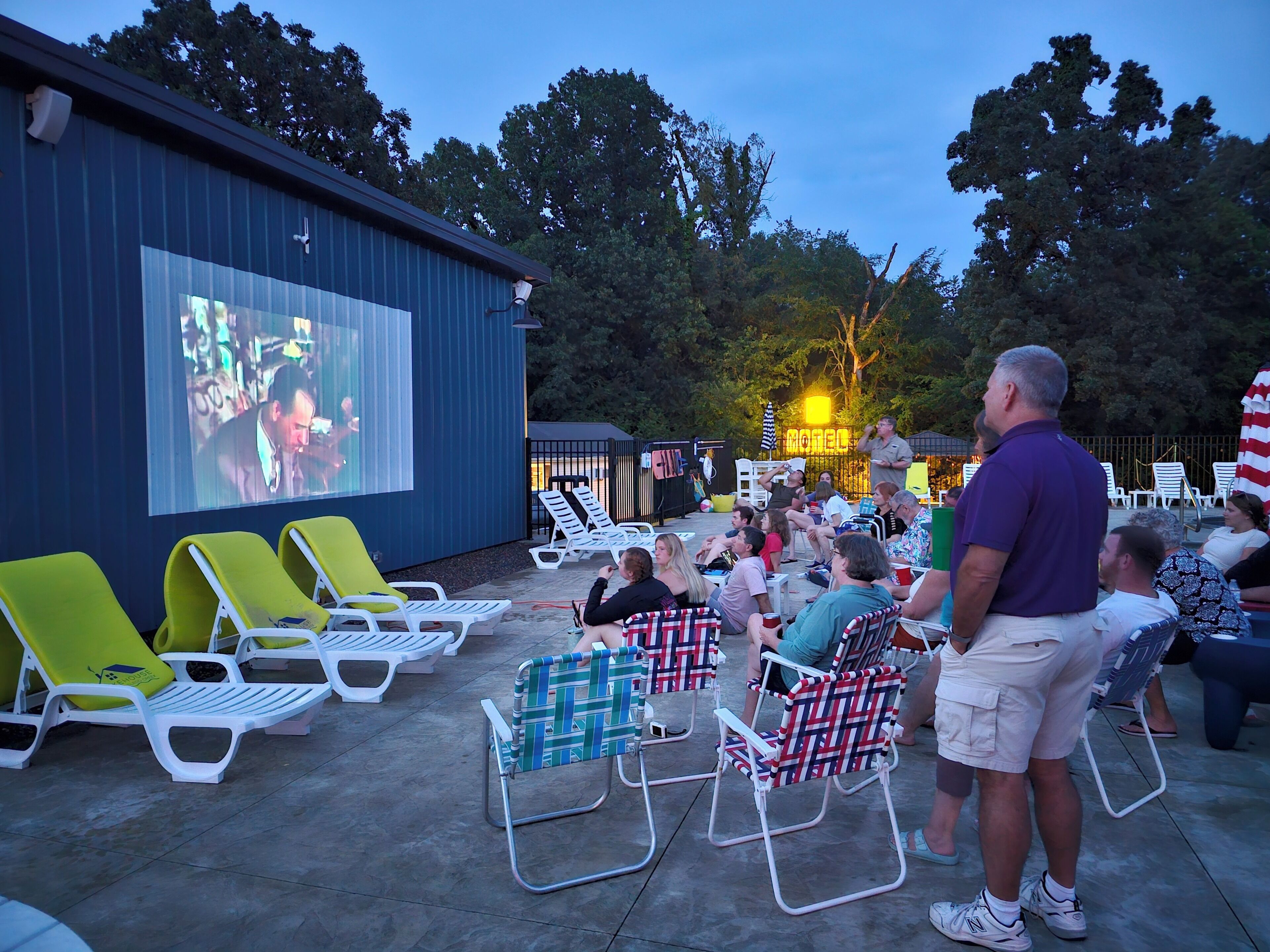 An outdoor movie screening area with a large screen, reclining chairs, and a building in the background.