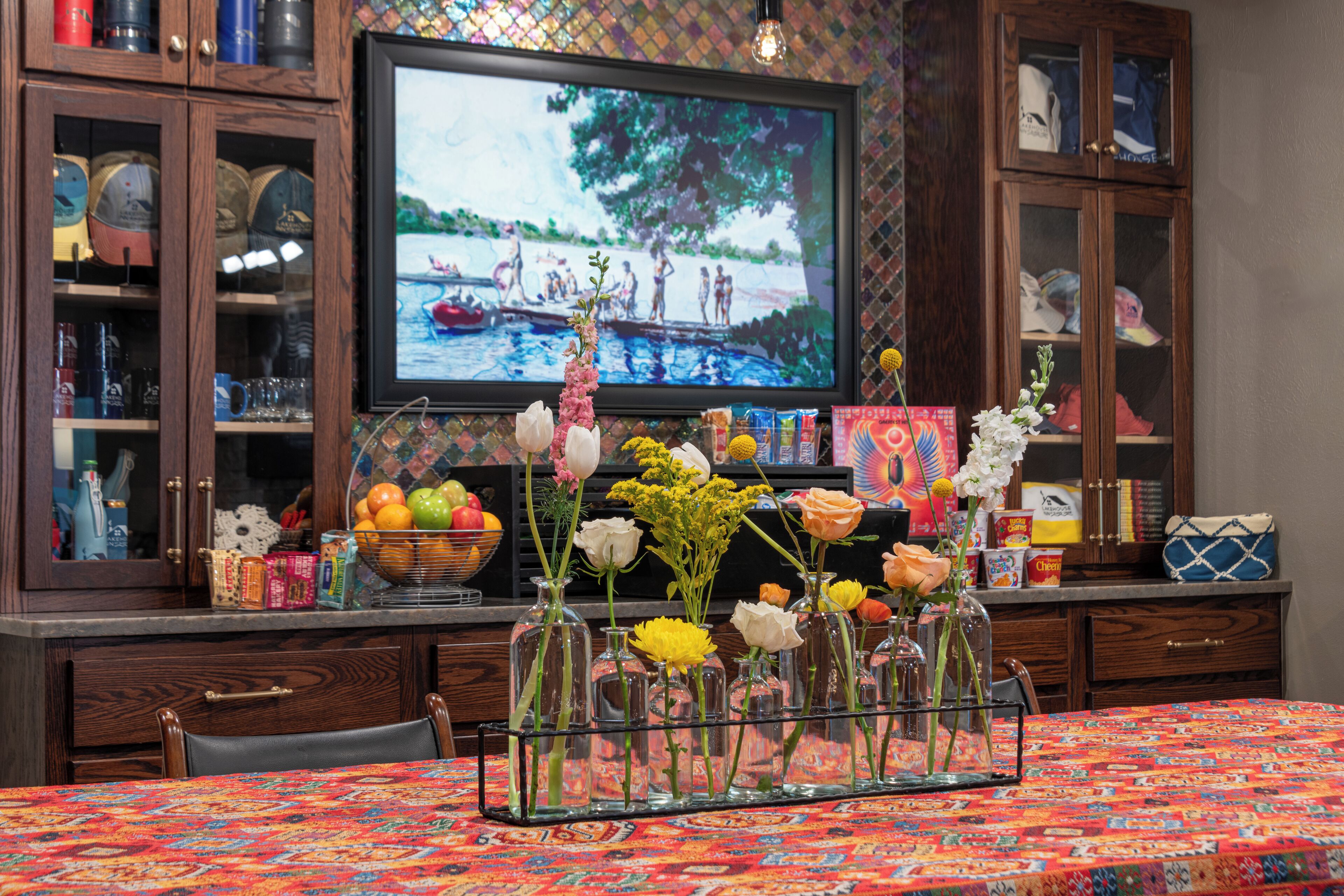 A dining area with a table covered by a colorful tablecloth, a television displaying a nature scene, and various decorative items including vases with flowers and a basket of fruits.
