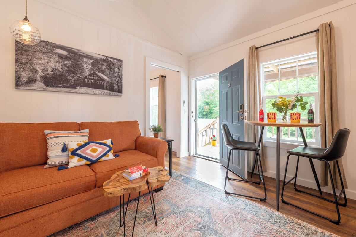 Ein Wohnzimmer mit Sofa, Couchtisch und Sesseln. An der Wand hängt ein Gemälde, eine Tür führt nach draußen und ein Fenster mit Vorhängen.