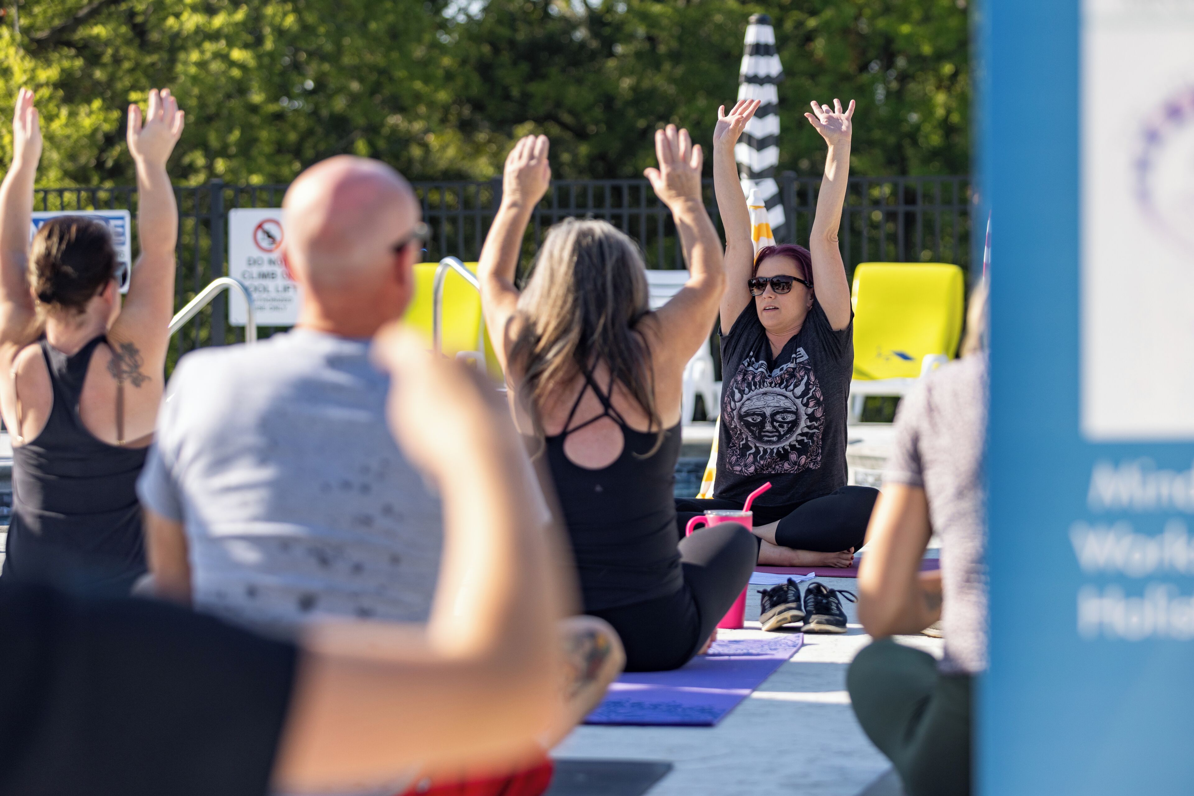 A group of people sitting on yoga mats outdoors, with trees and a fence in the background.