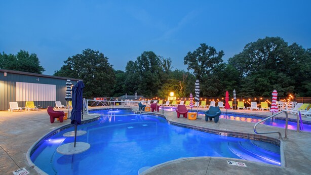 Une piscine dont le bord est illuminé de lumière bleue, entourée de chaises longues et de parasols aux couleurs vives, avec des arbres en arrière-plan.