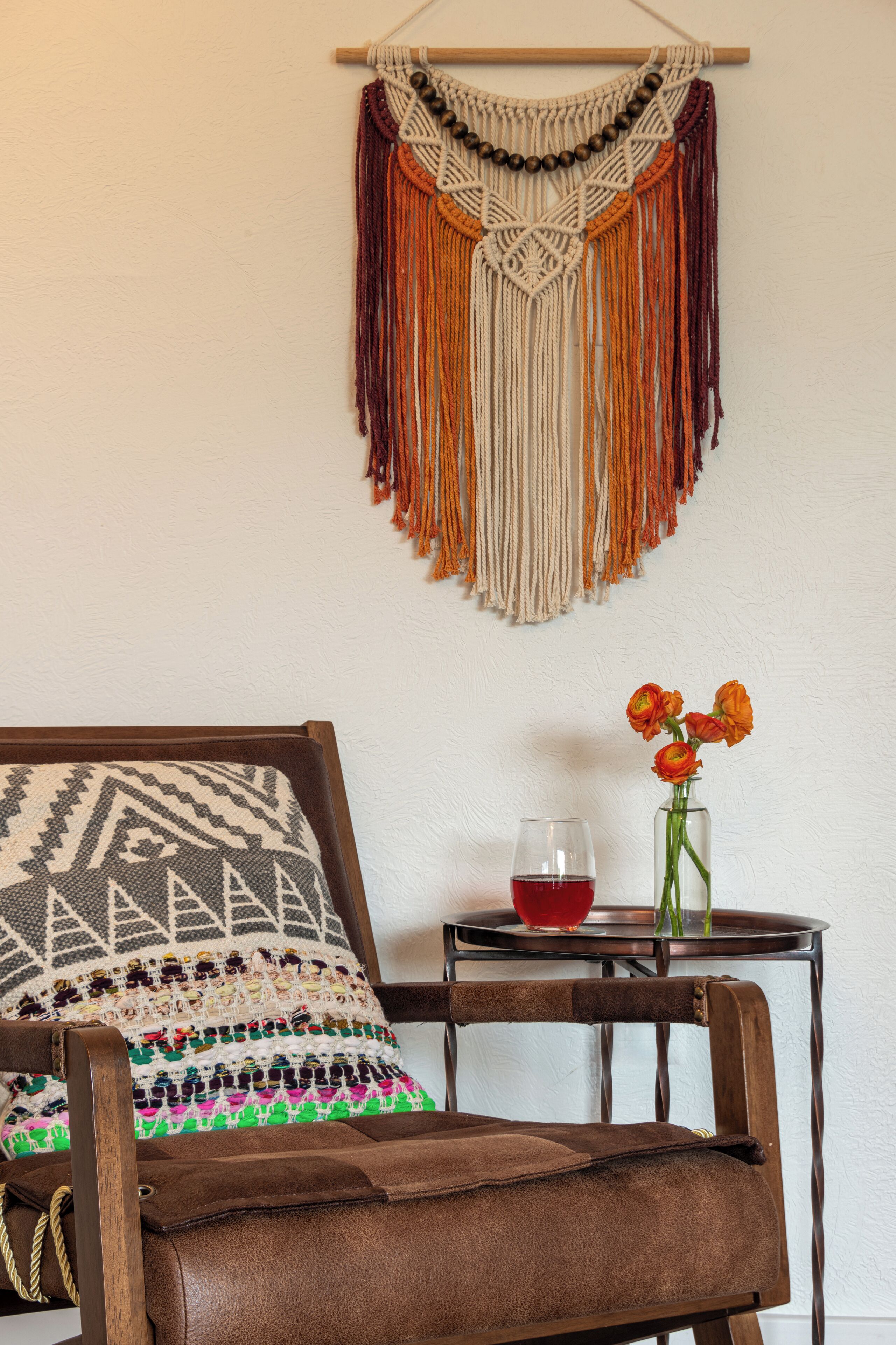 Een stoel met een gestreept kussen, een macramé wanddecoratie en een vaas met bloemen op een bijzettafeltje.