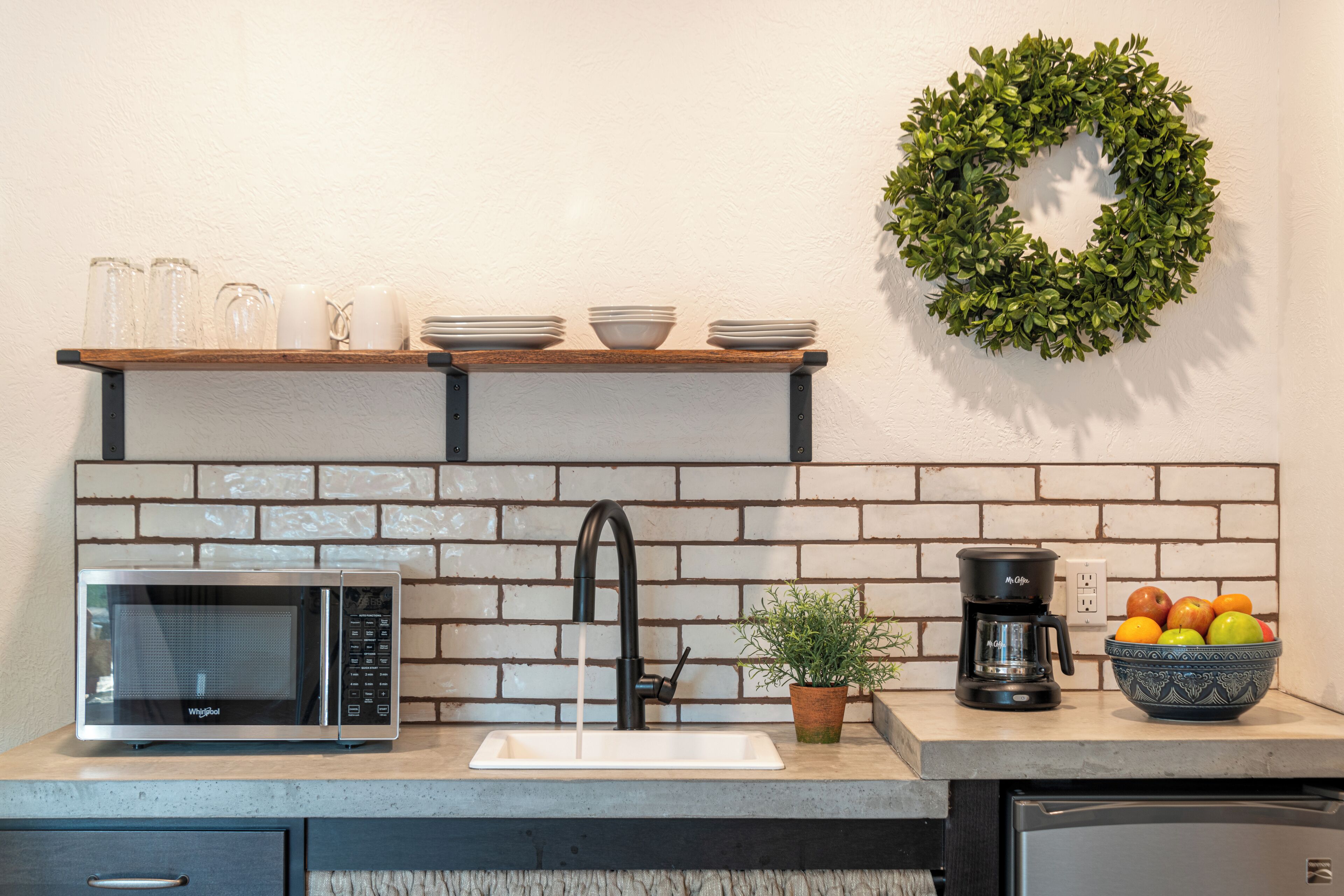 Una cocina con microondas, lavamanos, cafetera y un tazón de fruta.