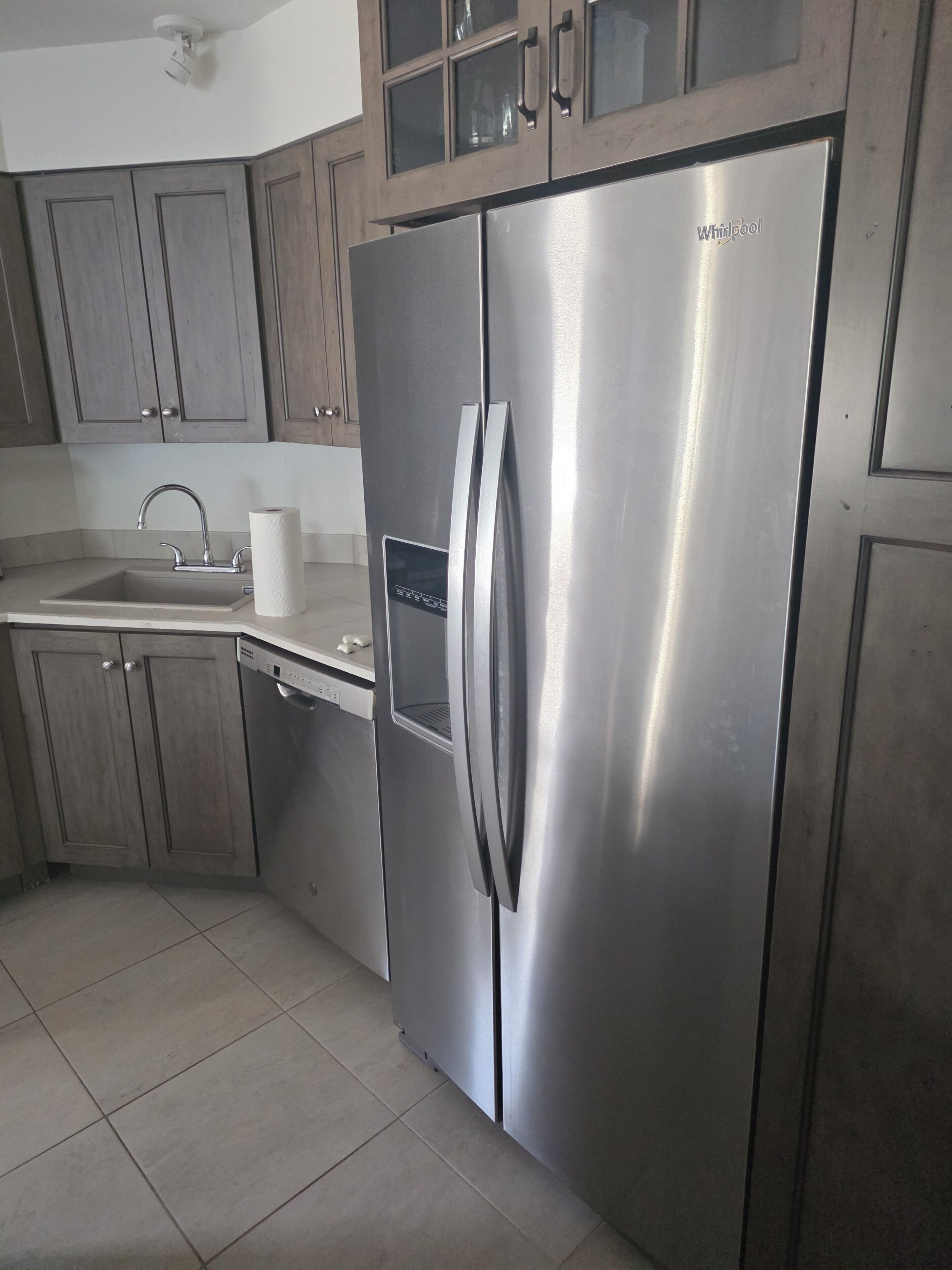 A stainless steel Whirlpool refrigerator in a kitchen with wooden cabinets and a tiled floor.