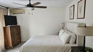 Un dormitorio con una cama, un ventilador de techo, un televisor sobre un mueble de madera, una mesita de noche con una lámpara, y dos cuadros en la pared.