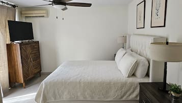 A bedroom with a bed, a ceiling fan, a TV on a wooden cabinet, a bedside table with a lamp, and two framed pictures on the wall.