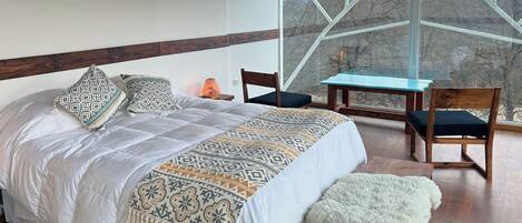 Un dormitorio con una cama grande, una silla, una mesita y vistas al exterior a través de un ventanal.