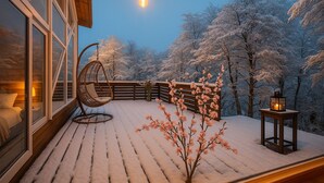 Een sneeuwbedekt balkon met een hangstoel, een tafeltje met een lantaarn, en uitzicht op met vorst bedekte bomen.