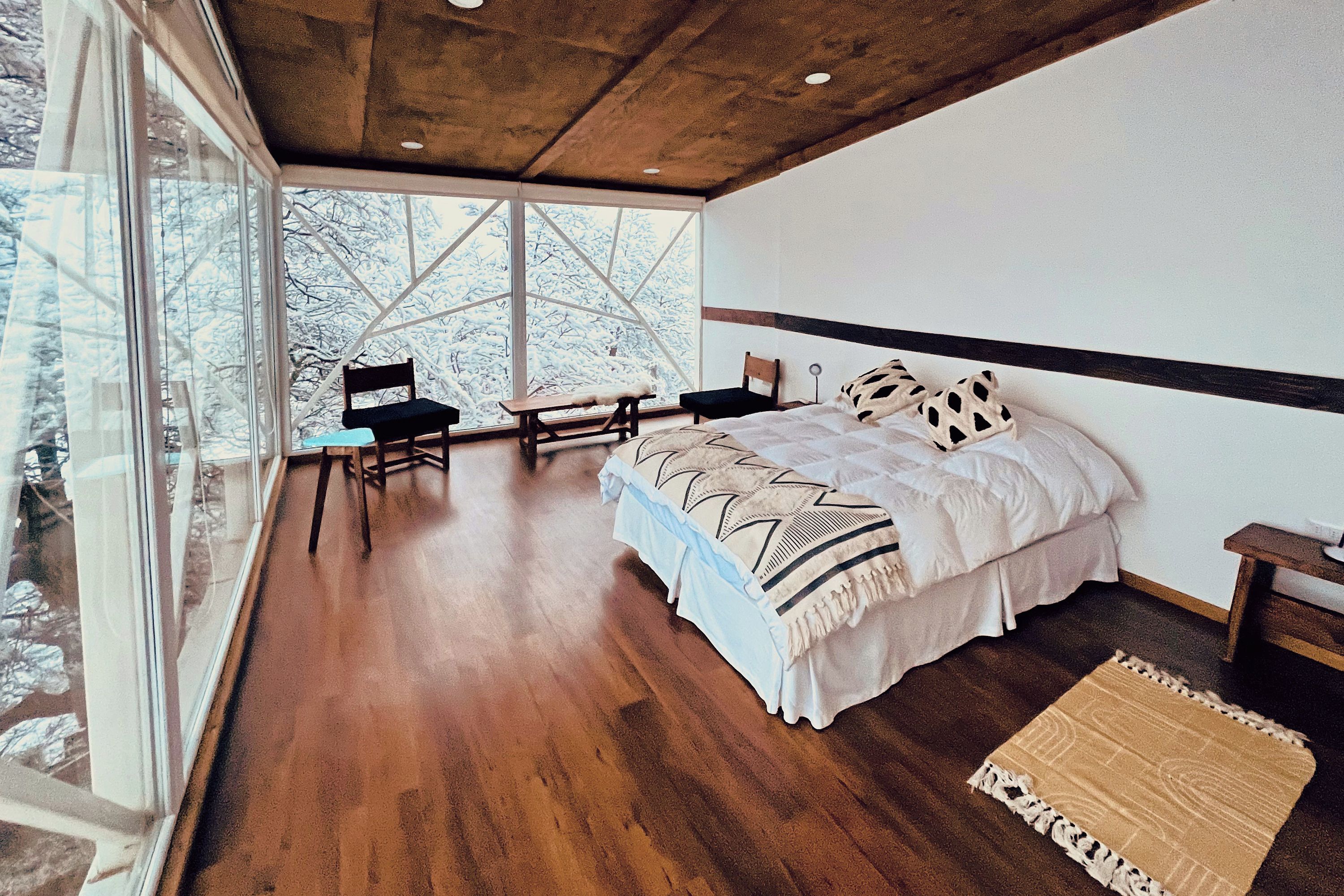 Ein Schlafzimmer mit einem großen Bett, Holzboden, Blick auf verschneite Bäume durch ein Fenster und einem kleinen Tisch mit Stuhl.