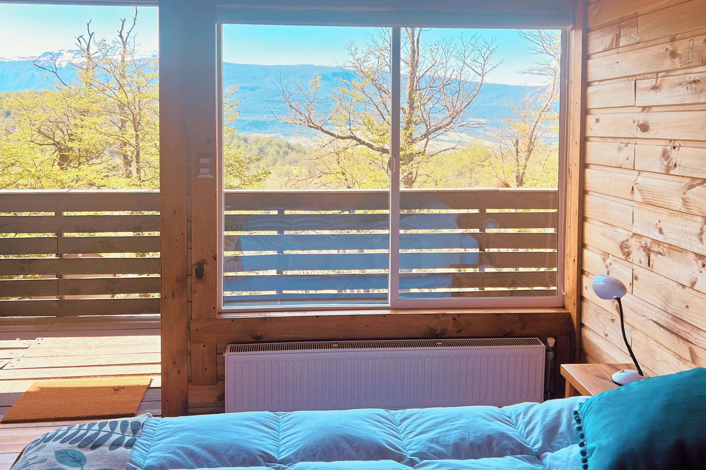 Una habitación con cama, ventana con vista a árboles y montañas, y piso de madera.