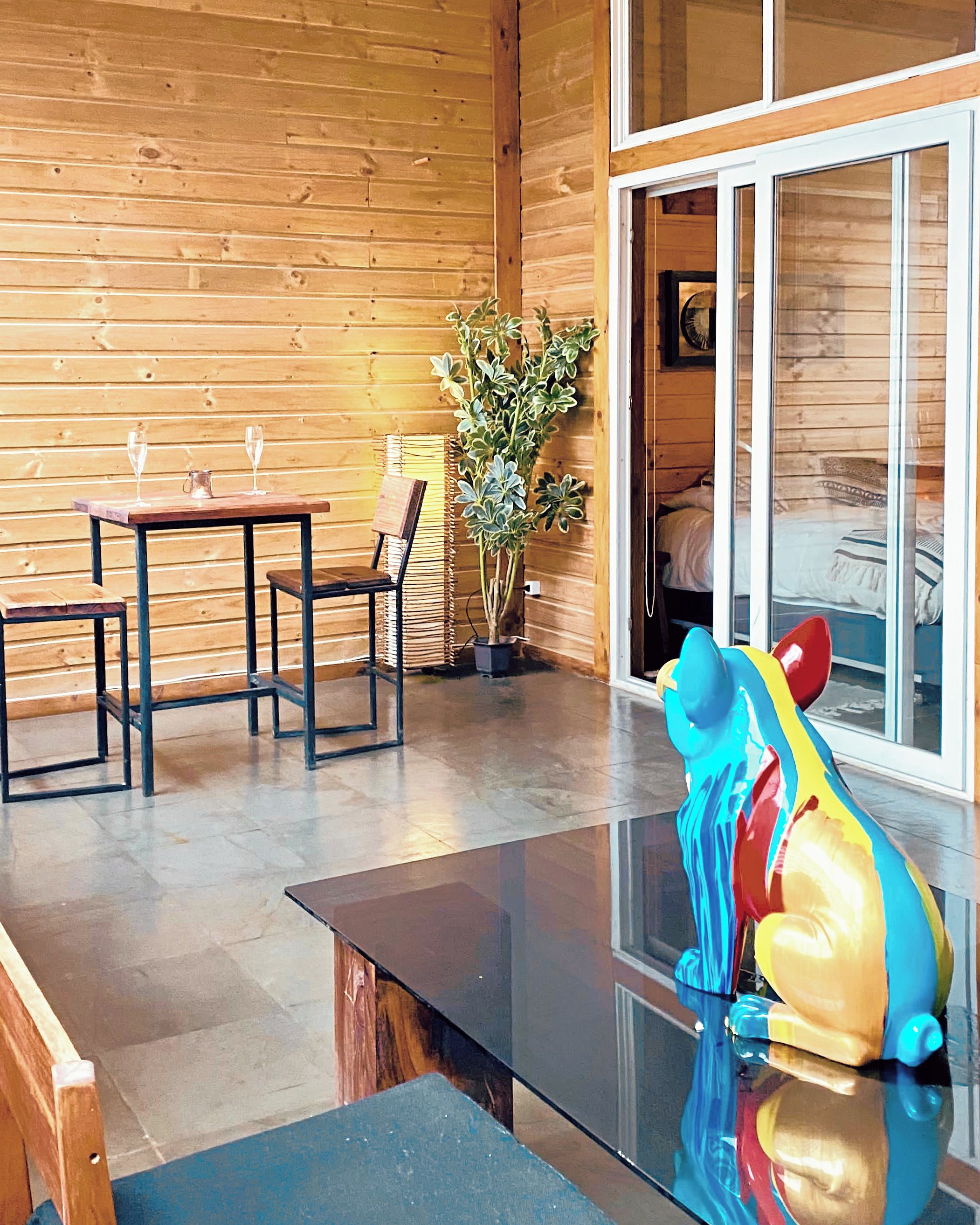 A wooden interior with a glass table, chairs, a potted plant, and a colorful sculpture.