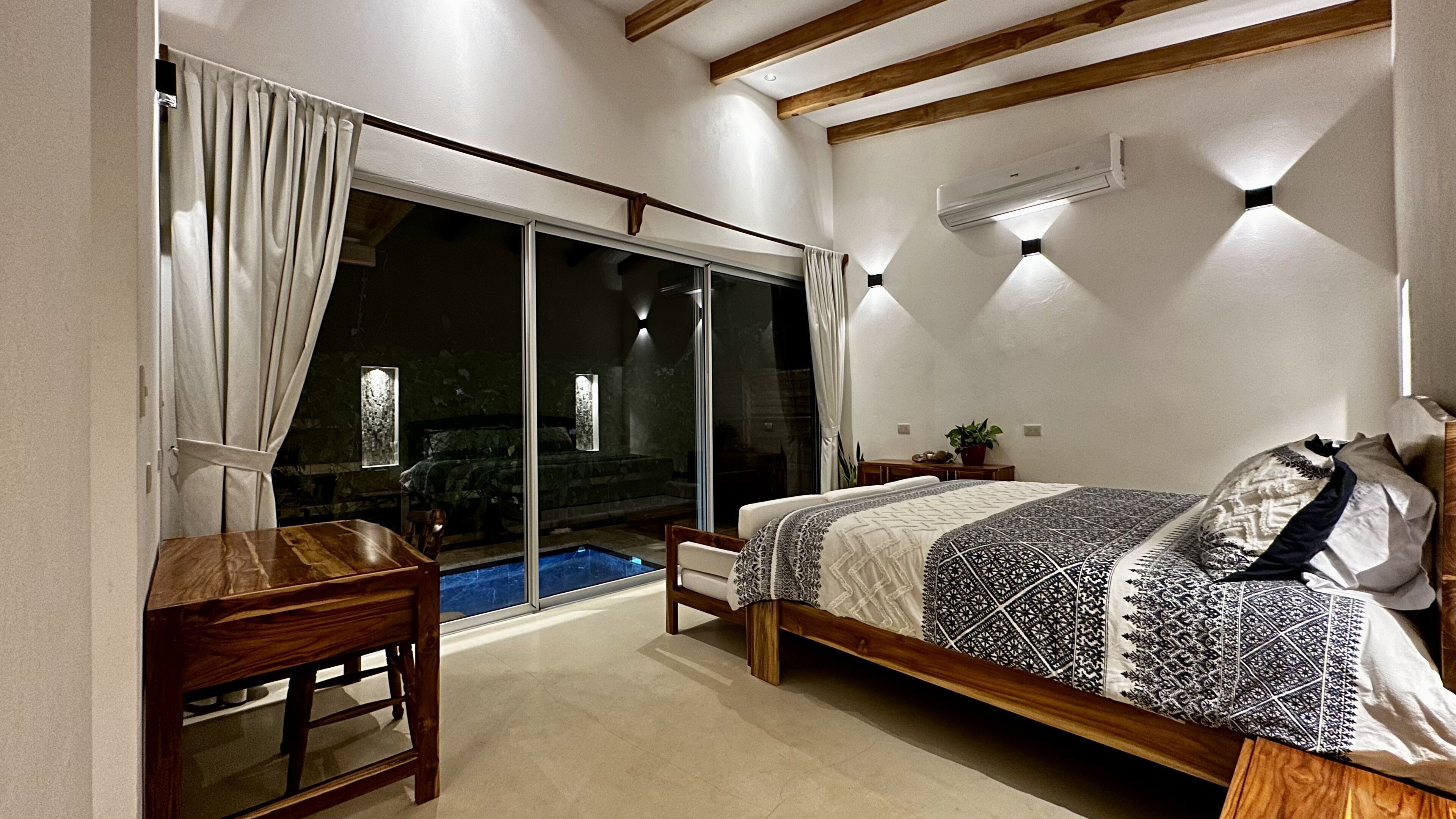 A bedroom with a large sliding glass door leading to a balcony, a bed with patterned bedding, a wooden nightstand, and an air conditioning unit on the wall.