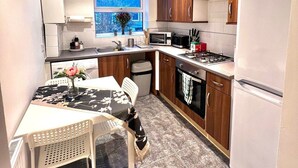 A compact kitchen with wooden cabinets, a white refrigerator, a microwave, and a stove. There is a window with a view of trees and a small table with a chair. - BAMBU (London)