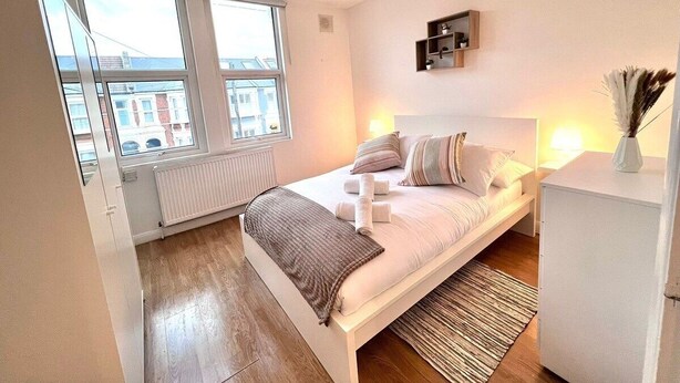 A modern bedroom with a large bed, a bedside table, a radiator, and a window with a view of buildings. - BAMBU (London)