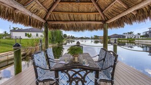 Un espacio de estar al aire libre cubierto con techo de paja, con vistas a un cuerpo de agua tranquilo y una zona residencial.