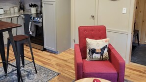 Un espace de vie compact comprenant une cuisine, un coin repas avec une table et des chaises, un fauteuil rouge agrémenté d’un coussin décoratif, et une table ronde sur laquelle se trouvent des pommes et une petite boîte.