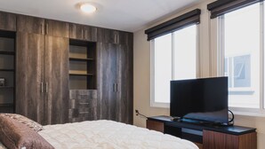 A bedroom with a bed, a TV on a stand, a window, and built-in wooden shelving.