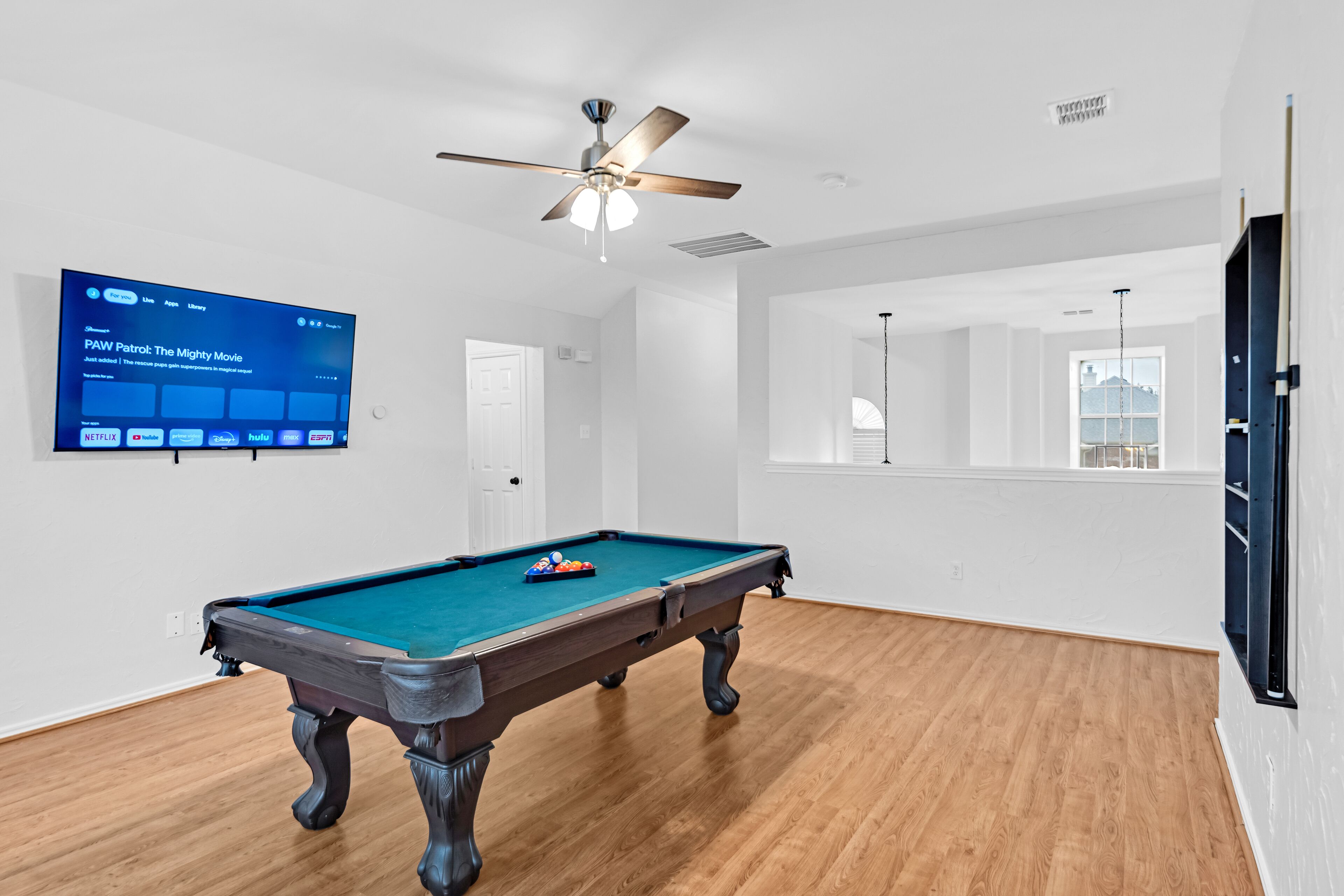 A pool table in a room with a ceiling fan, a mounted TV displaying a movie, and a large window.