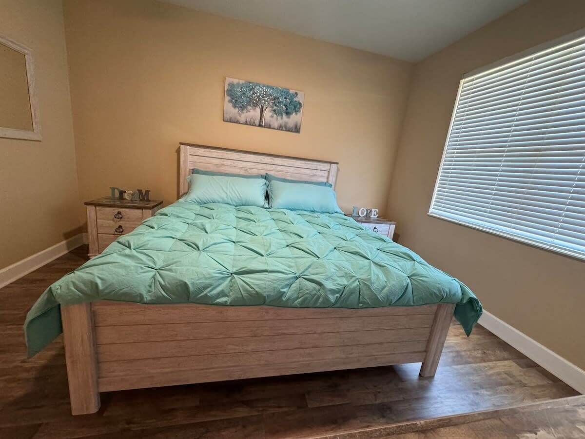 Un dormitorio con cama de madera, mesita de noche, cómoda, un cuadro en la pared y una ventana con persianas.