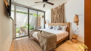 Quarto com cama, ventilador de teto, TV fixada na parede e porta de vidro deslizante que dá para área externa com plantas.