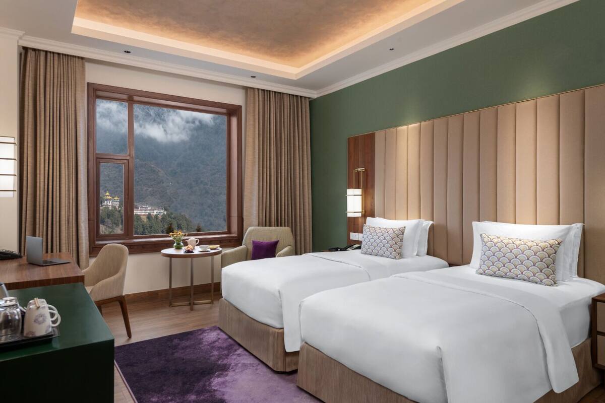 A hotel room with two beds, a desk with a laptop, a chair, a small table with a vase and teapot, and a view of a mountain.