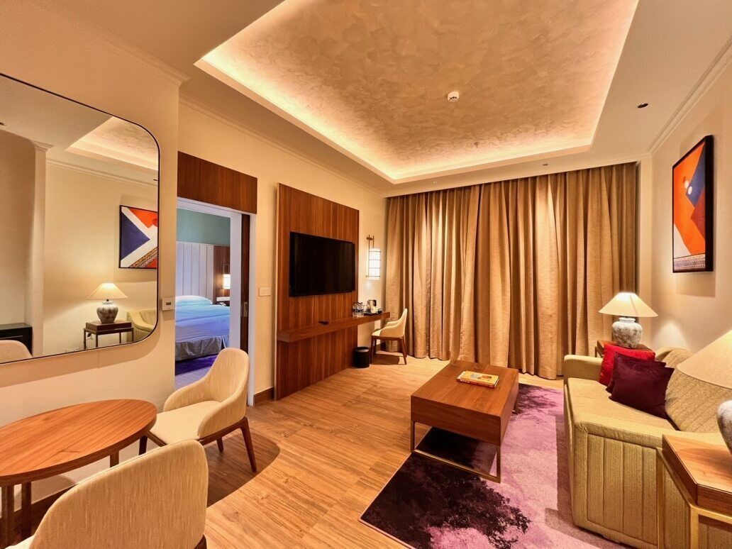A modern hotel room with a wooden floor, a flat-screen TV, a sofa, a dining table, and a large mirror.