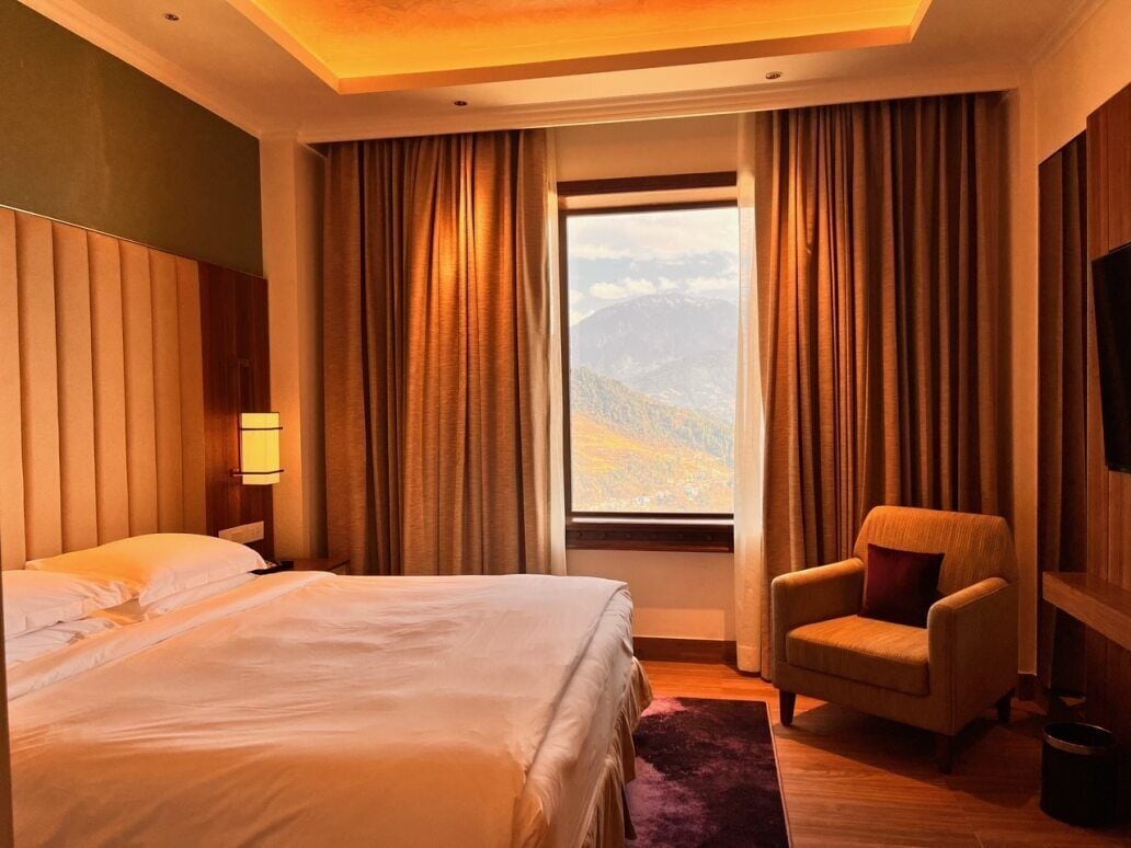 Habitación de hotel con una cama grande, una silla, una mesita de noche y vistas a las montañas a través de una ventana.