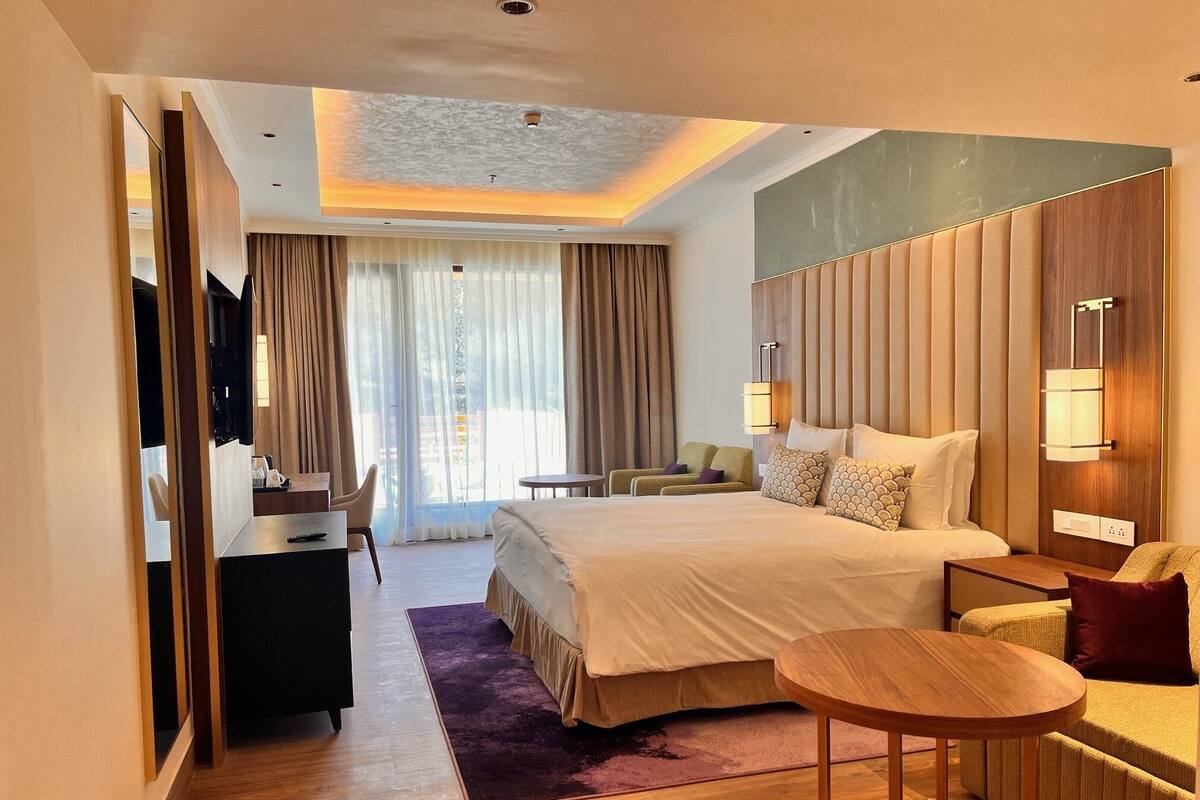 A modern hotel room with a large bed, a desk, a chair, a small table, and a TV.