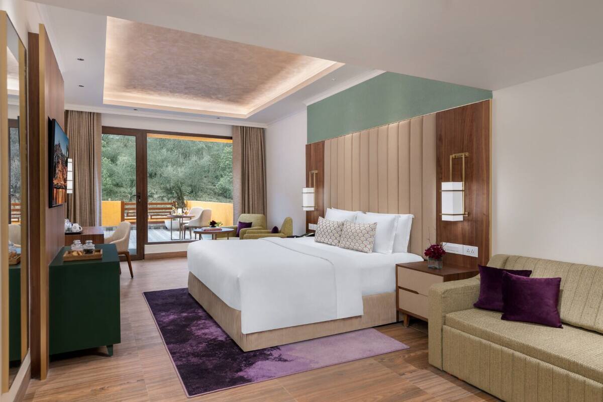 A modern hotel room with a large bed, a sofa, a desk, and a balcony with a view of trees.