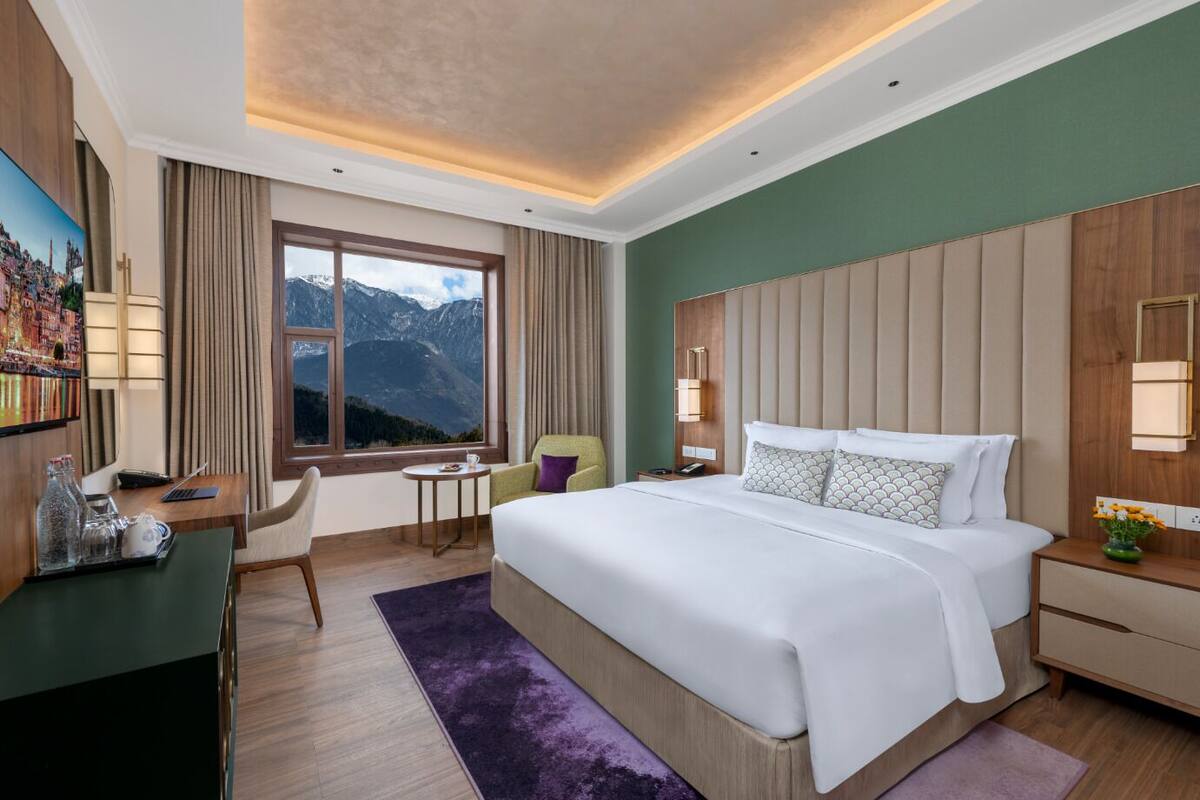 A hotel room with a large bed, a desk with a chair, a small table, and a window with a mountain view.