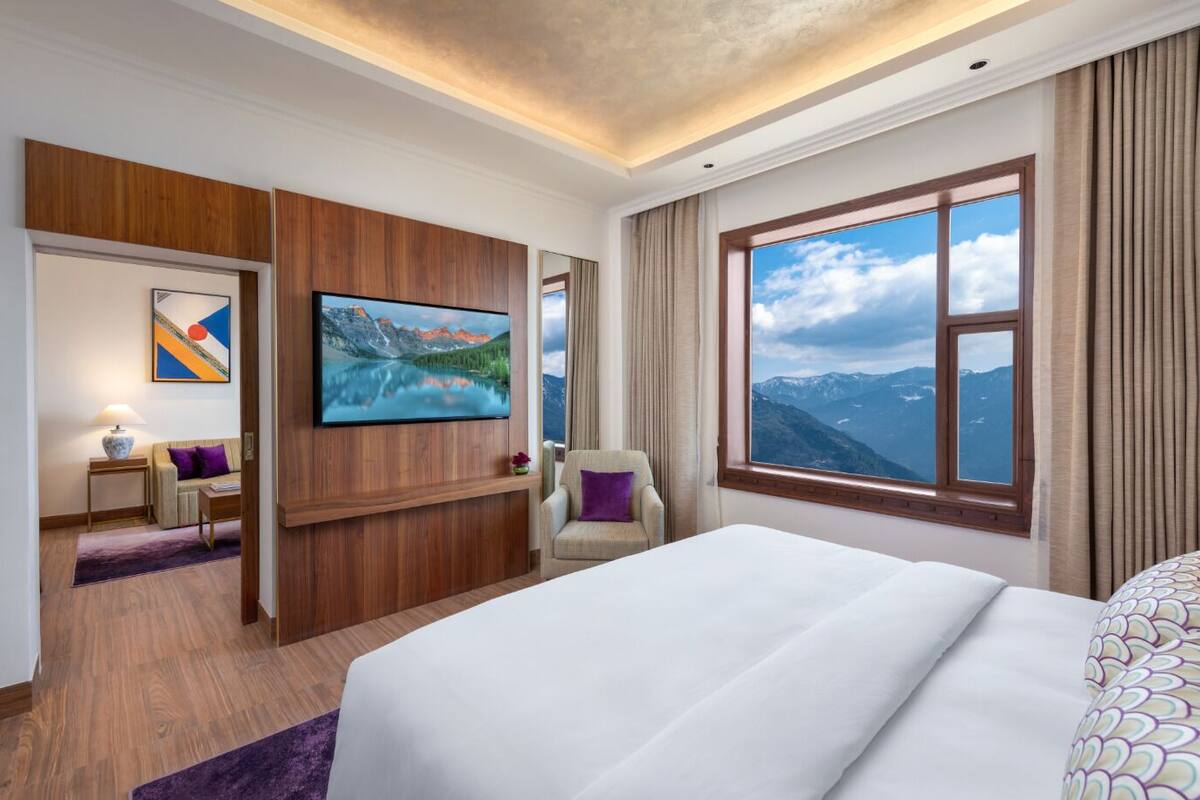 A hotel room with a large bed, a TV mounted on the wall, a chair, a small table, and a window with a mountain view.