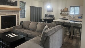 Un salon moderne avec un canapé d’angle, une cheminée et un coin repas, la cuisine étant visible au fond.