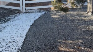 Un chemin de gravier mène à une clôture blanche, avec des arbres et un bâtiment en arrière-plan.