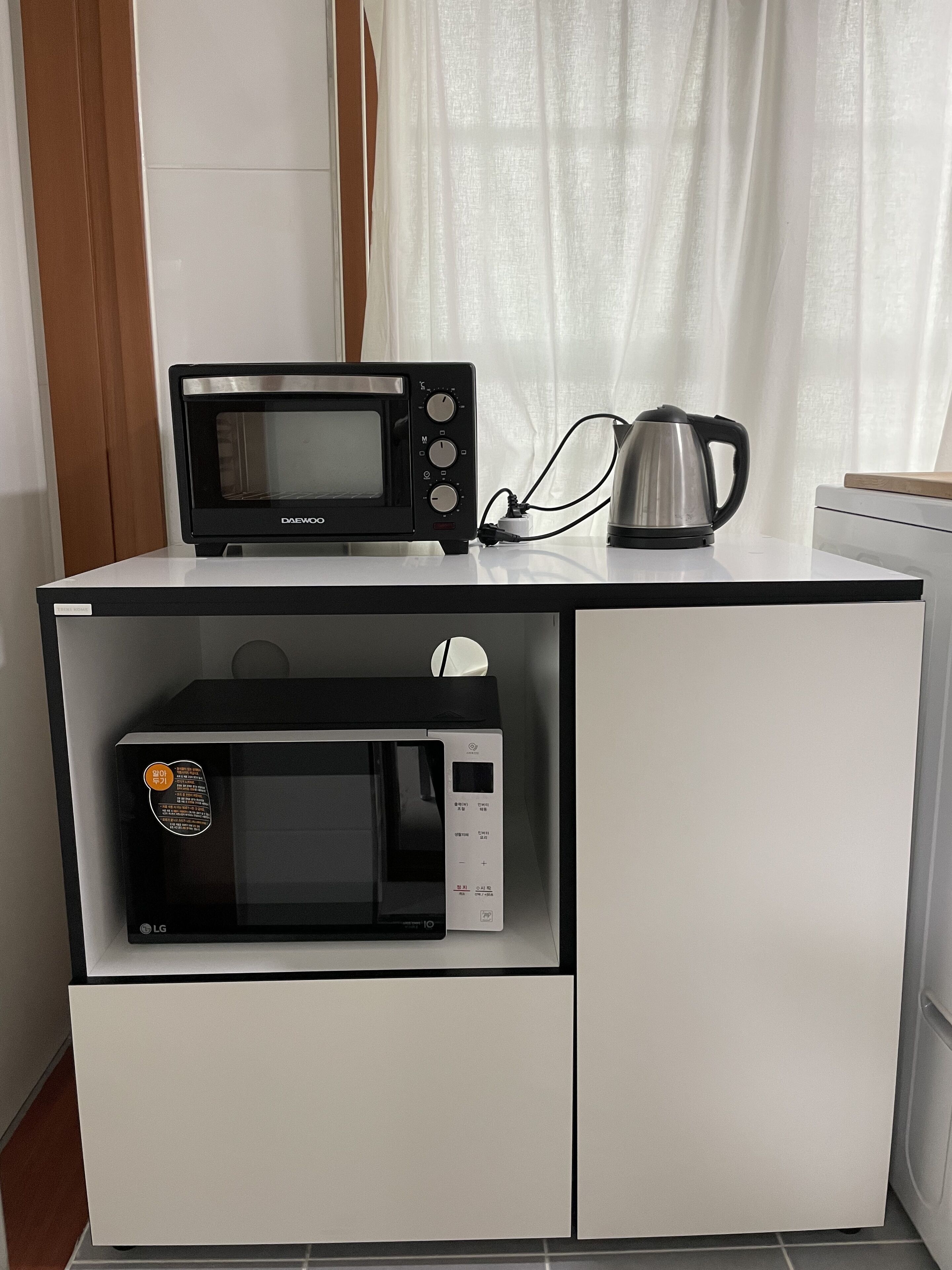 Eine Küchenzeile mit einer schwarzen Daewoo-Mikrowelle auf einem weißen Schrank, einer weißen LG-Mikrowelle darunter und einem Edelstahl-Wasserkocher auf der Arbeitsplatte.