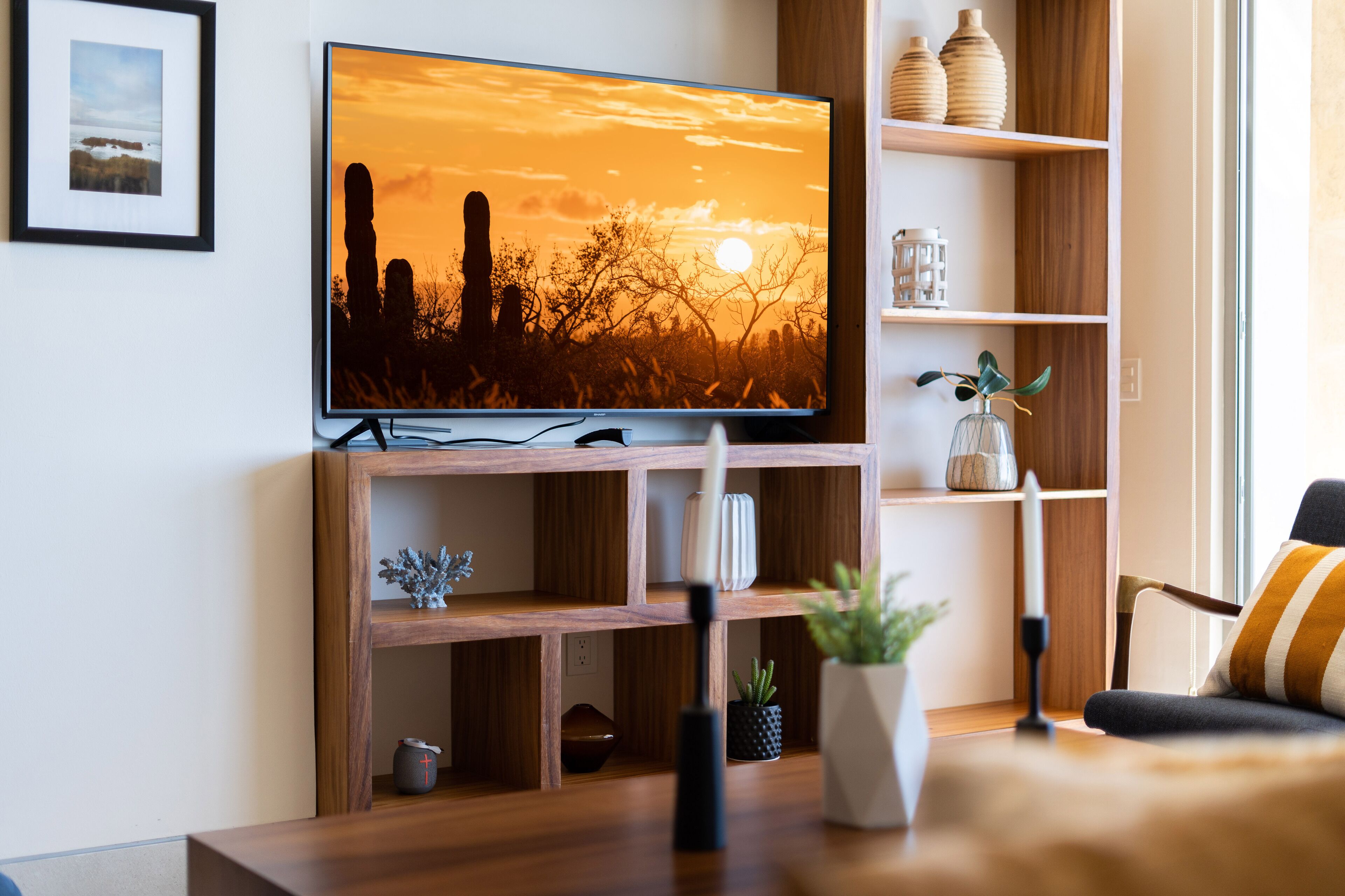 Una sala de estar moderna con un televisor de pantalla plana mostrando una escena de atardecer, estanterías de madera con objetos decorativos y una zona de estar acogedora.