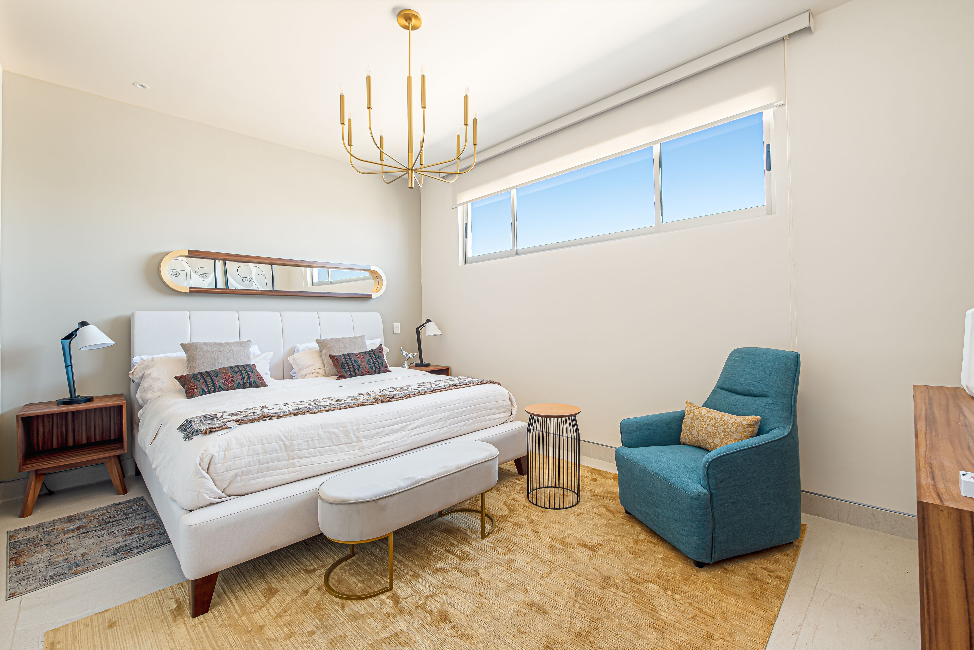 Une chambre à coucher moderne avec un grand lit, un fauteuil bleu, une petite table ronde et une grande fenêtre.