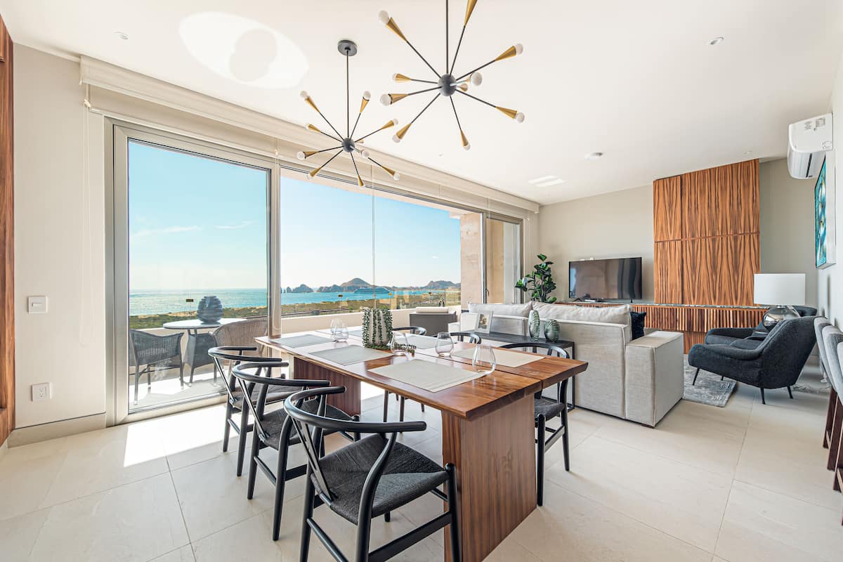 Una cocina moderna con una isla de madera, un comedor con sillas y un amplio ventanal corredizo que da a un balcón con vistas al océano.