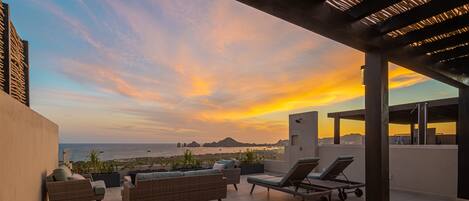A rooftop terrace with wicker furniture, a sun lounger, and a view of the ocean at sunset.