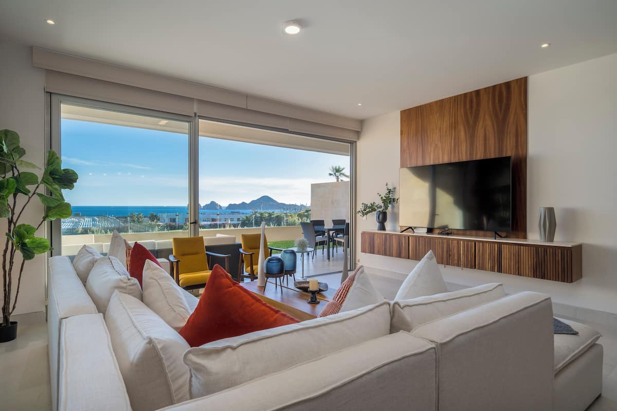 Una sala de estar moderna con un sofá grande, un televisor instalado en un panel de madera, y un balcón con vista al océano y montañas.