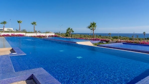 Una gran piscina azul cristalina con bordes de azulejos, rodeada de palmeras y flores rosadas.