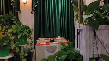 Espaço de refeições com uma mesa posta para dois, cercado por plantas em vasos e um fundo com cortina verde.