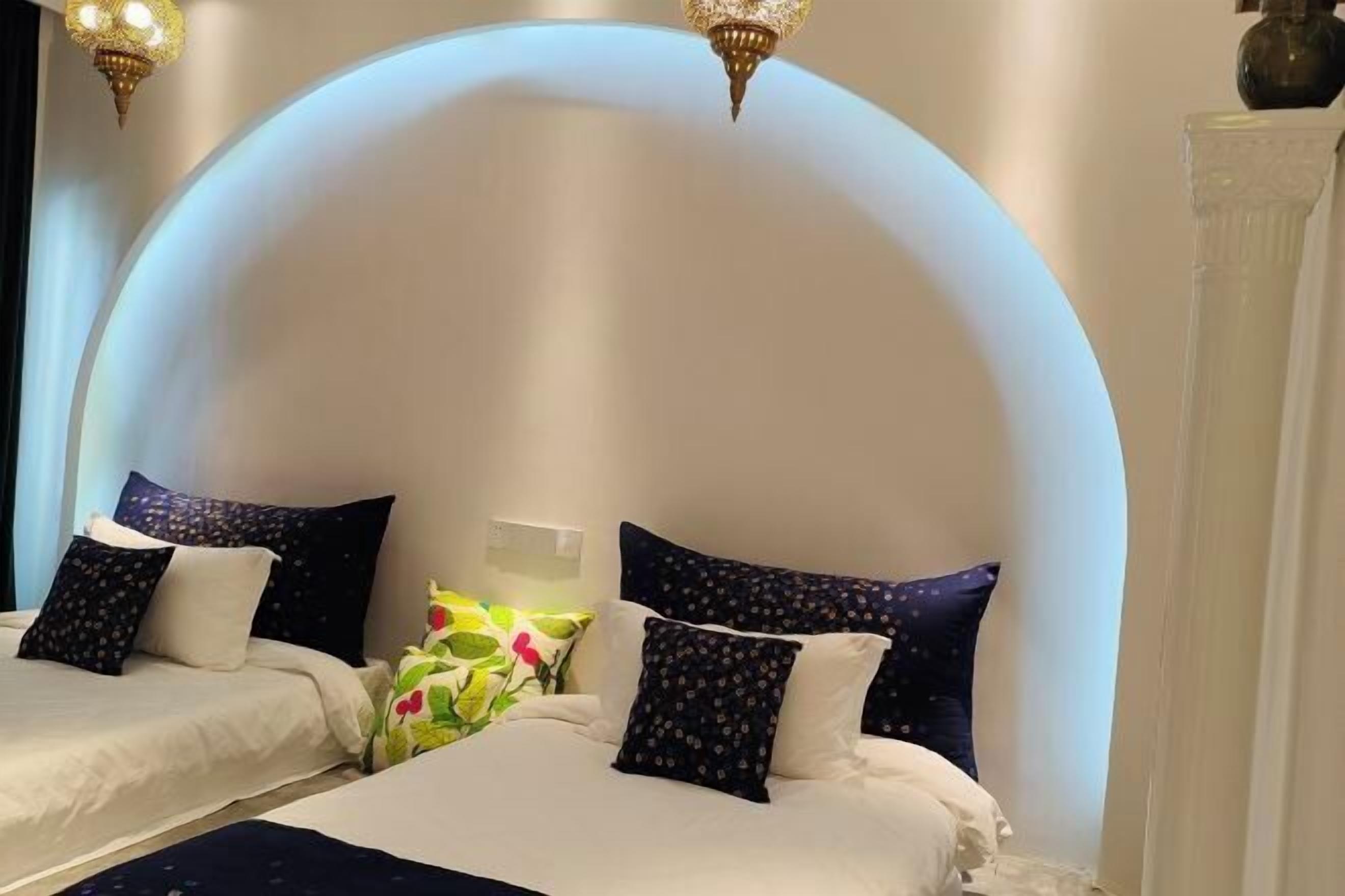 Habitación de hotel con dos camas, cada una con una colcha blanca y almohadas oscuras con estampado. Entre las camas hay una pequeña mesa con un jarrón de flores.