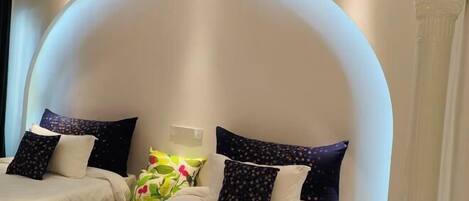 Habitación de hotel con dos camas, cada una con una colcha blanca y almohadas oscuras con estampado. Entre las camas hay una pequeña mesa con un jarrón de flores.