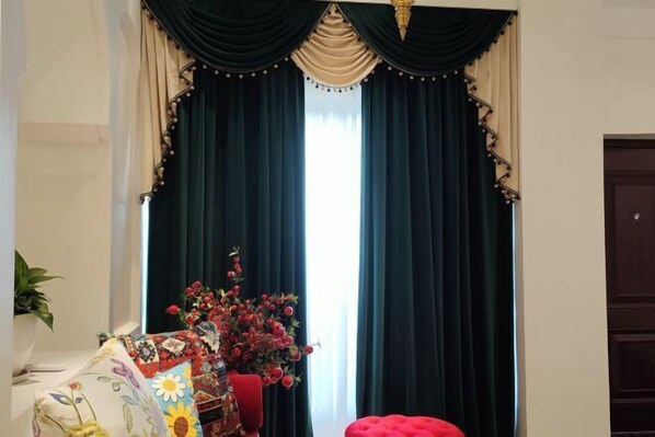 A room with dark green curtains, a floral arrangement, and a red ottoman.