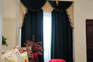 A room with dark green curtains, a floral arrangement, and a red ottoman.