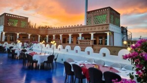 An outdoor dining area with tables and chairs, a covered walkway, and a building with patterned walls. - Silk Road Lodges (Turpan)