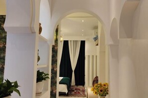 A narrow hallway with an arched doorway leading to a room with a bed, a rug, and a vase of sunflowers.