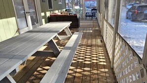 Et overdækket udendørs siddeområde med picnicborde og en stol, en træterrasse og udsigt til en rød bygning gennem vinduerne.